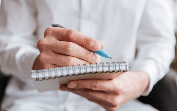 Close-up of a person's hands holding a pen and writing in a spiral notebook.