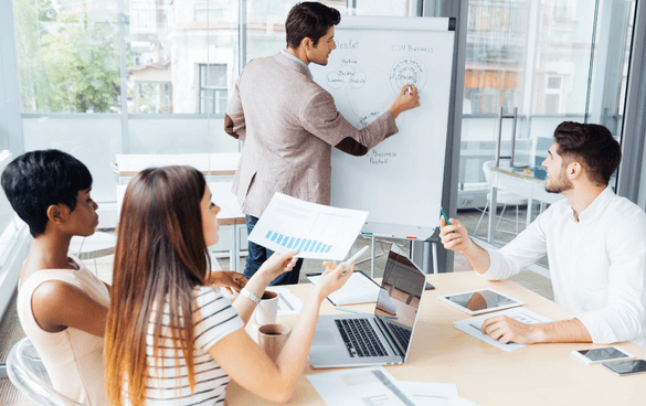 Four professionals in a meeting with one man presenting CRM basics on a whiteboard to three colleagues who are sitting at a table with laptops and documents, in a bright office environment.