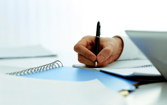 Close-up of a person's hand writing in a notebook with a pen, with a laptop and papers on the desk.