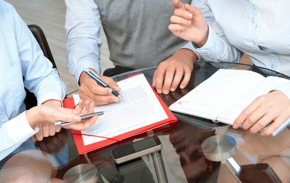 Three individuals engaged in a business meeting, with one pointing to a document, another holding a mobile phone, and a third with an open notebook on the table.