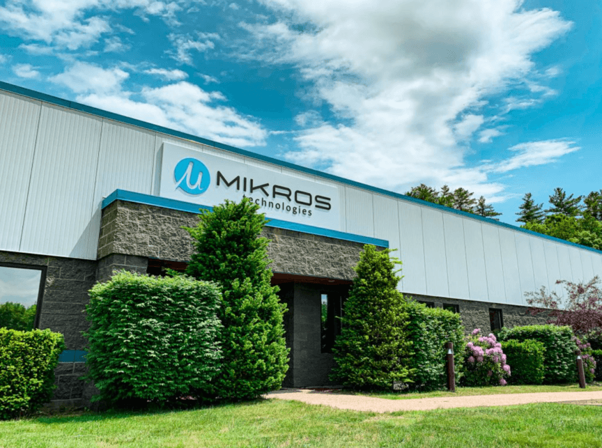 A single-story commercial building with 'Mikros Technologies' signage, surrounded by green shrubs and trees under a partly cloudy sky.