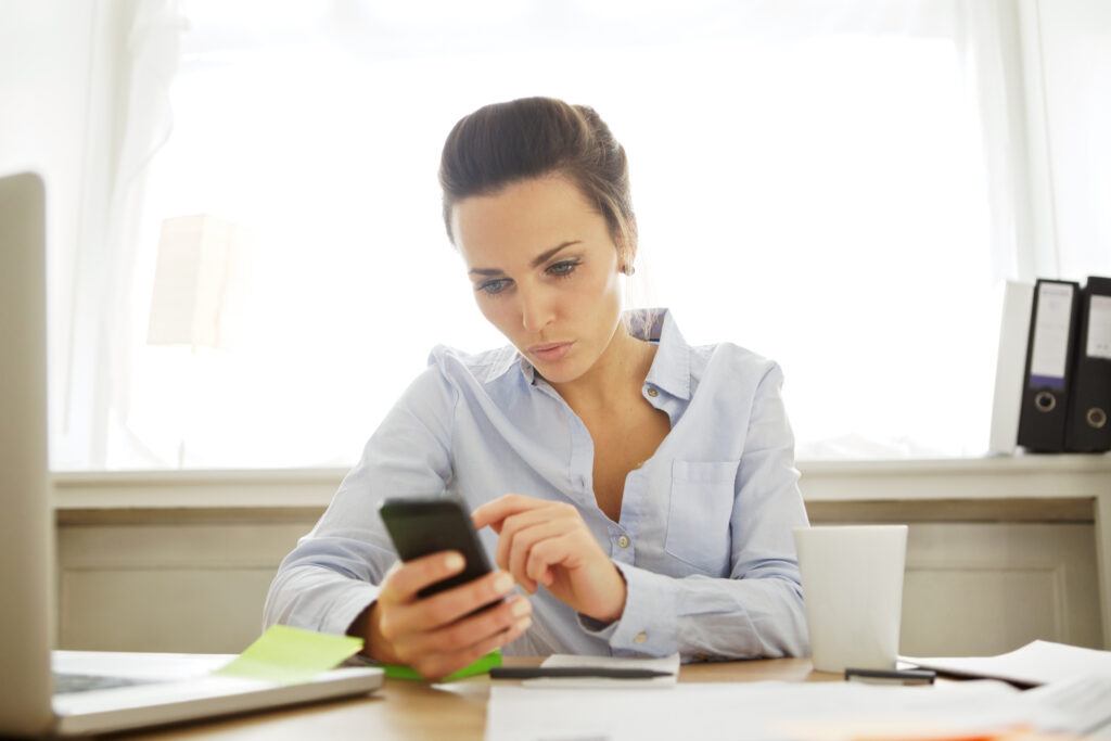 Young businesswoman in office with mobile phone sending a text message. Caucasian young woman sitting at the desk working. Female working in home office.