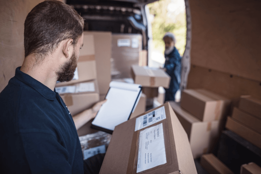 A delivery man inside a van checks a package against a clipboard while another person waits outside, surrounded by numerous packages.