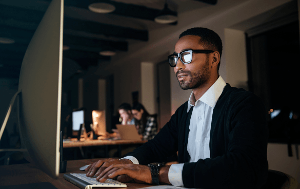 A focused man in glasses and business attire working on a computer in a modern office setting with colleagues in the background.