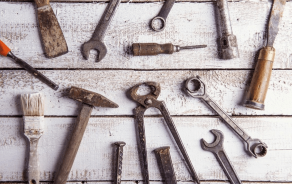 A variety of old, rusty tools such as a hammer, wrenches, screwdriver, paintbrush, chisel, nails, and screws laid out on a white wooden surface.