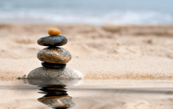 A stack of balanced stones with a reflection on water, set against a sandy beach and blurred ocean background.