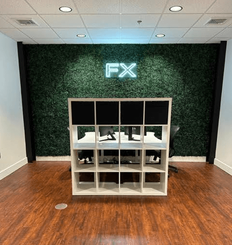 Modern office space with a greenery wall with illuminated 'FX' letters, a white cubicle with two workstations, each with dual monitors, keyboards, and mice on a wooden floor.