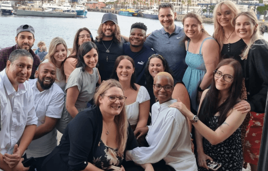 A diverse group of people smiling and posing together on a waterfront with boats in the background.