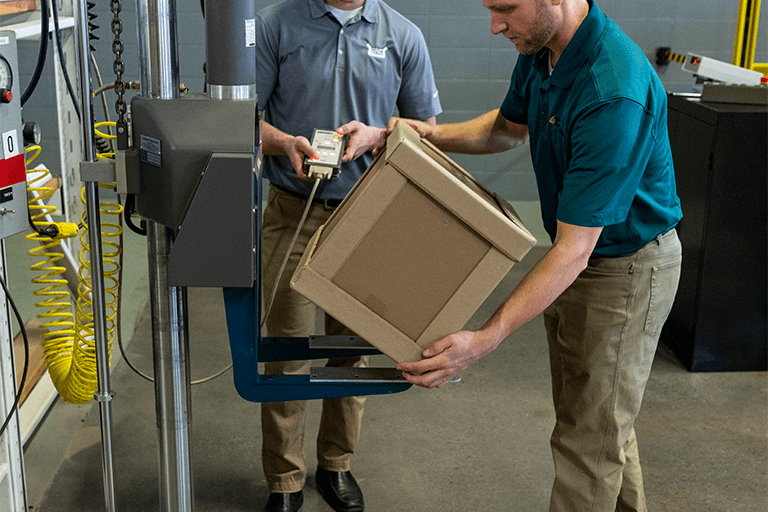 Two men in a workshop, one lifting a cardboard box onto a blue scale while the other holds a handheld device, possibly overseeing a quality control or shipping process.