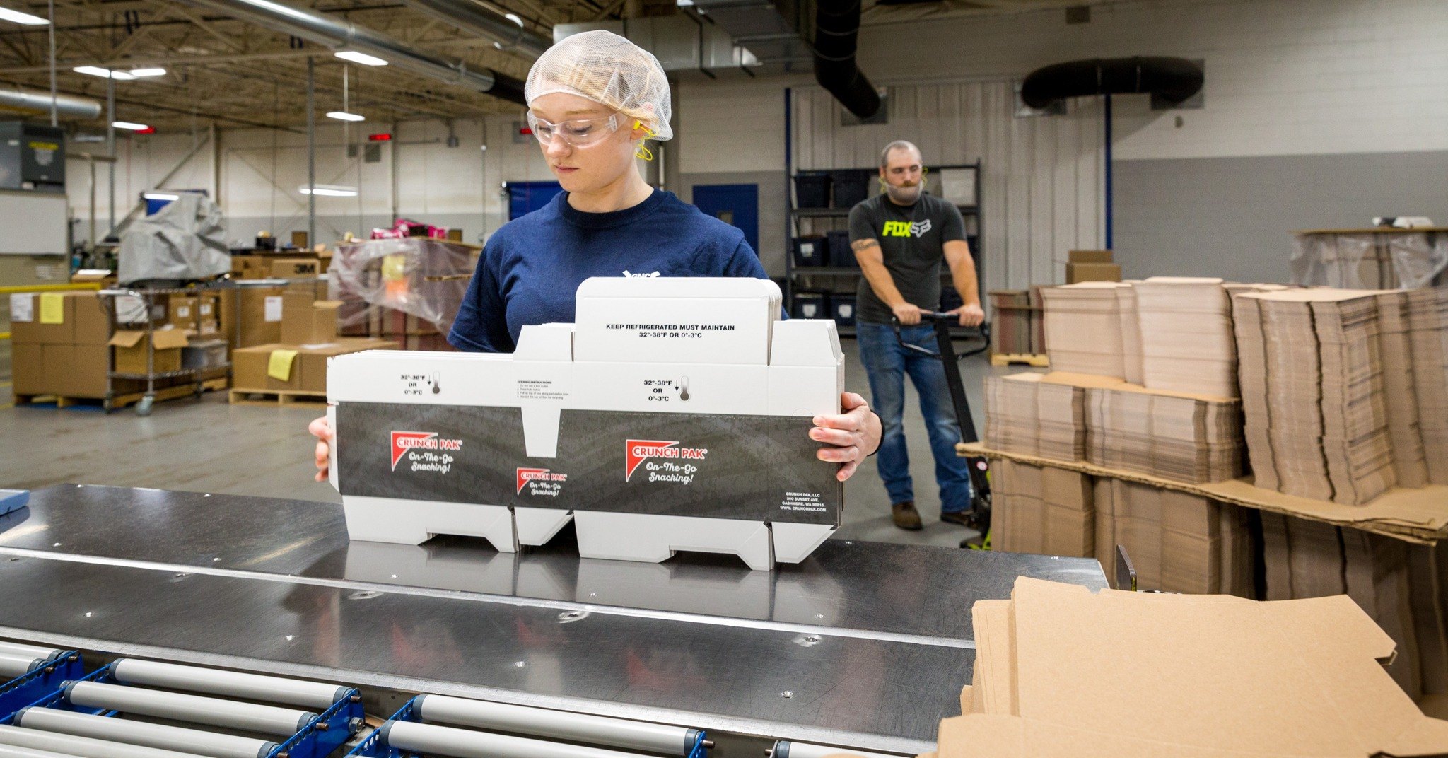 A woman in a factory wearing a hairnet and safety glasses holds an open cardboard box with refrigeration instructions, with a man and packaging materials in the background.