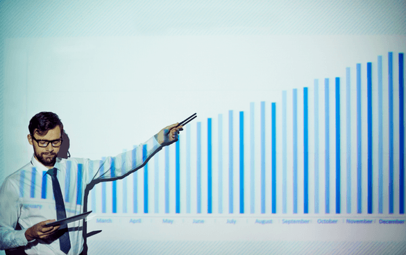 A man in a white shirt and tie presenting a monthly bar chart on a screen, using a pointer to highlight specific data while holding a tablet.