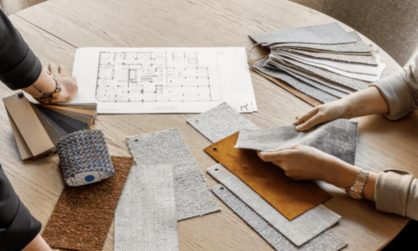 Two individuals collaborating over an interior design project with fabric samples and a floor plan on a wooden table.