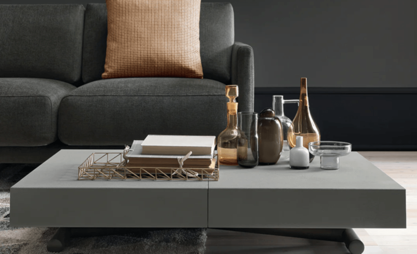 Modern living room with a dark grey sofa and a large grey coffee table displaying books, decorative glass vases, a gold-colored bottle, and a white candle.