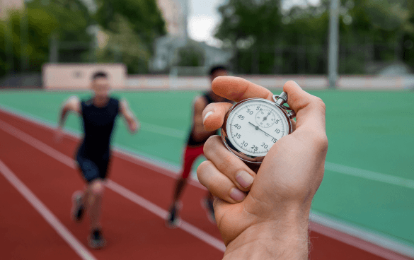 A hand holding a stopwatch with a focus on the timer, as two athletes sprint on a track in the background.