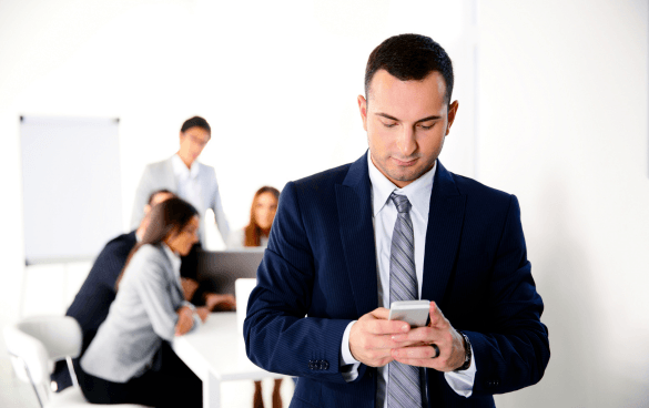 A man in a suit checking his smartphone in the foreground with a group of colleagues discussing in the background in an office setting.