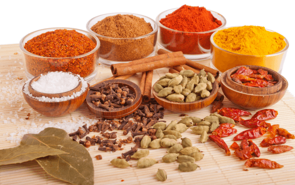 An assortment of spices and seasonings on a light surface, including bay leaves, salt, cloves, cinnamon, green cardamom, dried red chilies, and bowls of various ground spices like a spice blend, chili powder, and turmeric.