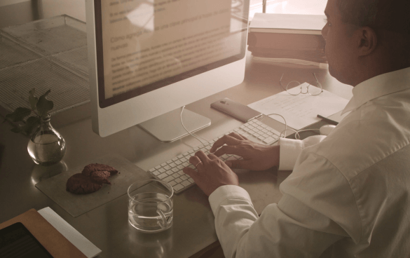 Person working on an iMac at a desk with a keyboard, a glass of water, cookies on a plate, a potted plant, and a book with a bookmark.