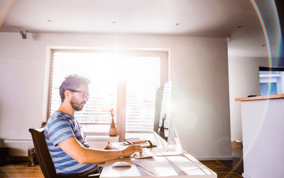 Man with glasses and a beard working on a desktop computer in a bright, modern room with sunlight shining through the window.