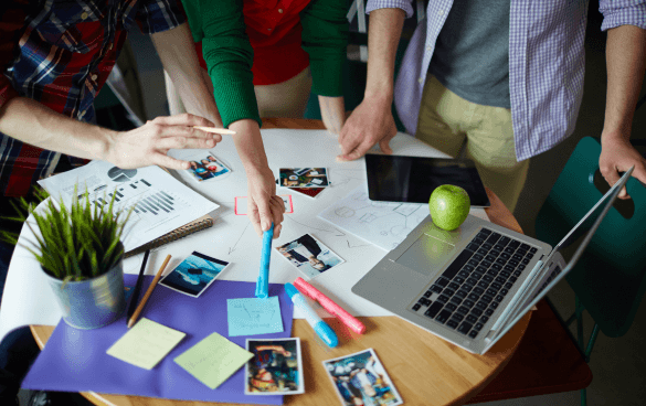 A group of people collaborating around a table with a laptop, tablet, photographs, sticky notes, markers, a green apple, and a small potted plant, indicating a creative work session.