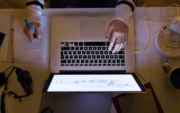 Person working at a desk with an open laptop displaying a document, while writing notes in a notebook and surrounded by papers and headphones.