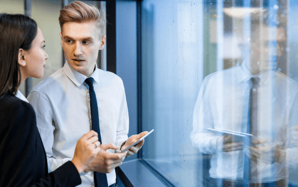 A man and a woman in business attire having a conversation next to a rain-spattered glass window, with the man holding a smartphone and looking at the woman.