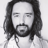 Portrait of a man with shoulder-length wavy hair and a beard, looking directly at the camera with a slight smile, wearing a white shirt with an open collar.