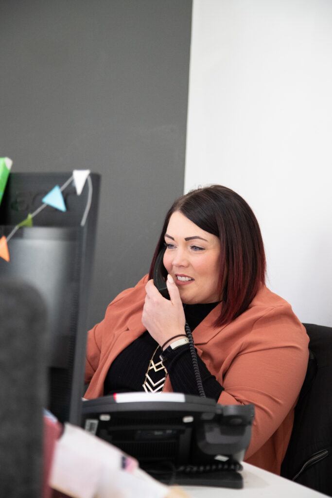 A woman in a peach blazer and black top speaking on a phone at her office desk, with a computer monitor and small decorative flags in the background.