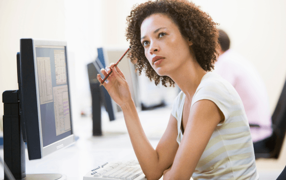 A thoughtful woman with curly hair holding a pencil to her chin while looking at a computer screen in an office setting.