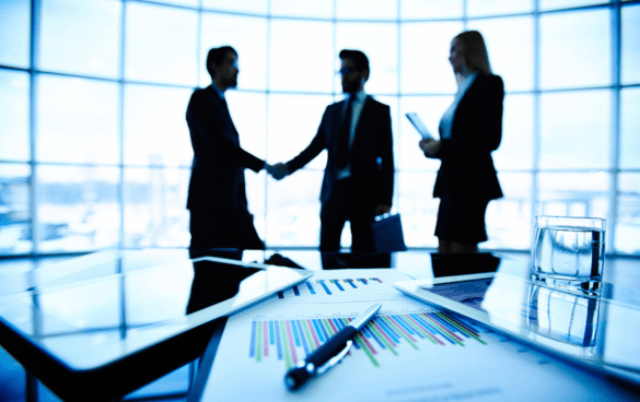 Two business professionals shaking hands in a well-lit office with another observing, in front of a conference table with documents, indicating a business meeting or agreement.