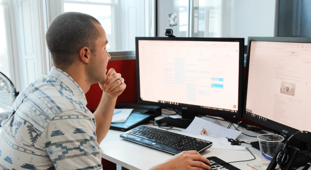 A man sitting at a desk working on a computer with two monitors, surrounded by office items like a keyboard, mouse, headset, and papers.