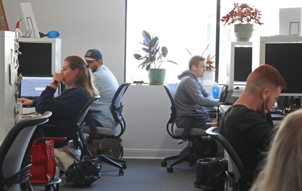 Individuals working in a modern office setting with computers, ergonomic chairs, and decorative plants, with one person wearing headphones.