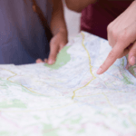 A person's finger pointing at a location on a paper road map while another person looks on.