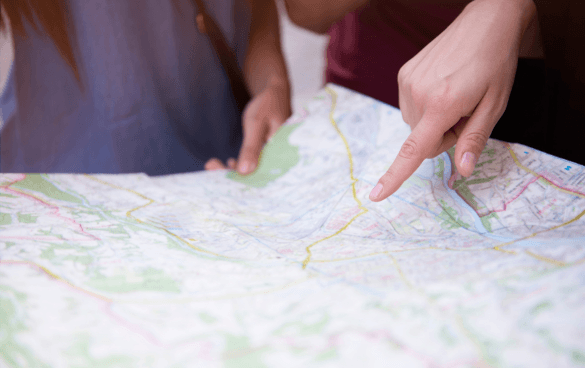 A person's finger pointing at a location on a paper road map while another person looks on.