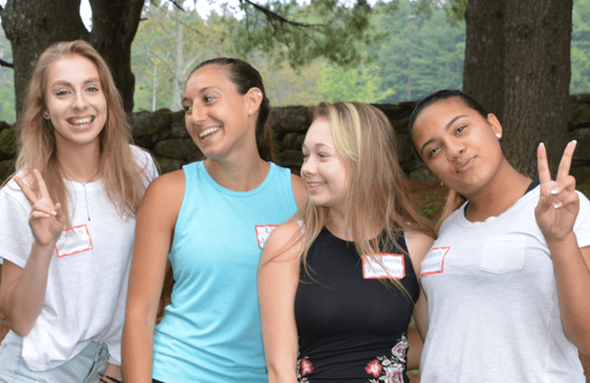 Four young women smiling and posing outdoors, two of them making peace signs, with trees and a stone wall in the background.