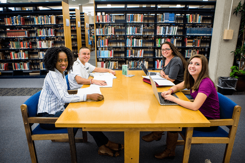 Four students sitting at a table in a library, two with laptops and two with papers, smiling at the camera.