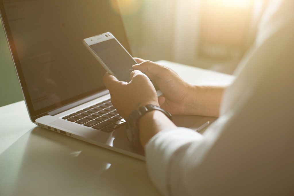 Silhouette of cropped shot of a young man working from home using smart phone and notebook computer, man's hands using smart phone in interior, man at his workplace using technology, flare light.