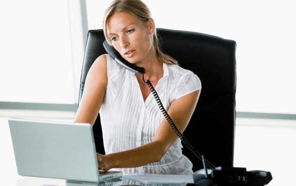 A woman multitasking in an office, using a laptop and talking on the phone while holding the receiver with her shoulder.