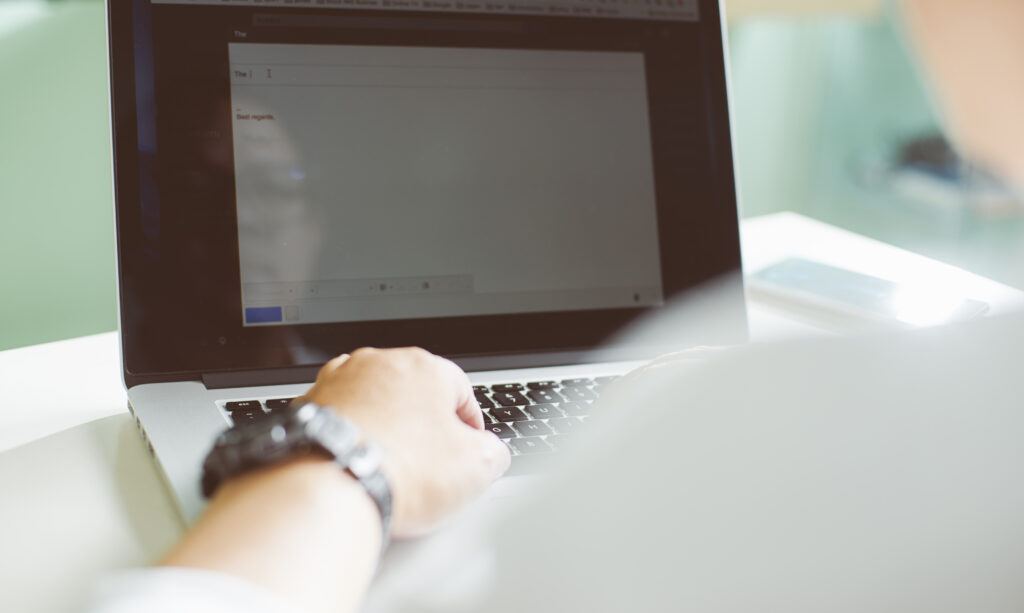 Person typing on a laptop with the start of a document on the screen, wearing a watch on their left wrist, with a smartphone lying next to the laptop.