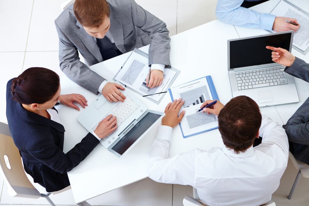 Four professionals in a meeting discussing data with a laptop, tablet, and printed charts on the table.