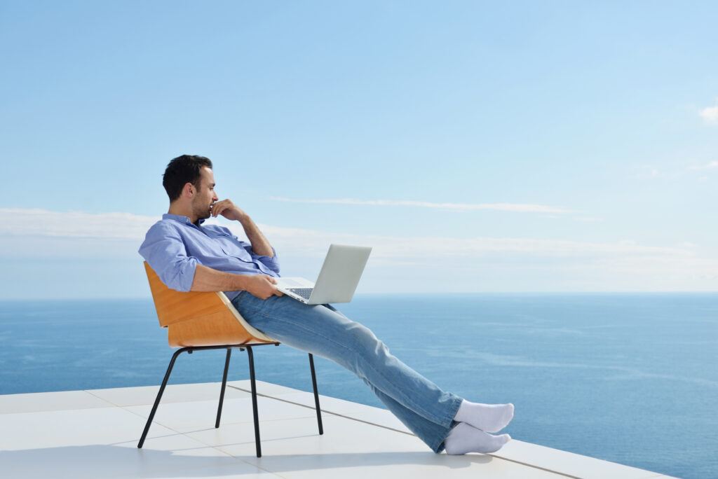 Man in casual attire working on a laptop while sitting on a chair on a balcony with a clear view of the sea and sky.