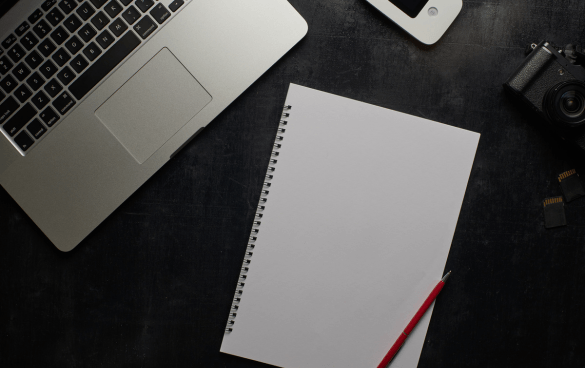 A workspace with a laptop, a blank spiral notebook with a red pencil, a camera, a smartphone, and two SD cards on a dark surface.