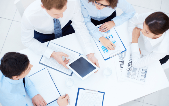 Bird's-eye view of five professionals in a meeting, with documents, a tablet, and a newspaper on the table, indicating a collaborative work environment.