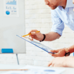 Two professionals reviewing a document with charts and graphs in the background, indicating a business or project discussion.
