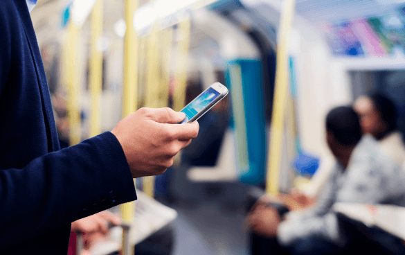 Close-up of a person's hand holding a smartphone inside a subway train, with other passengers in the background.