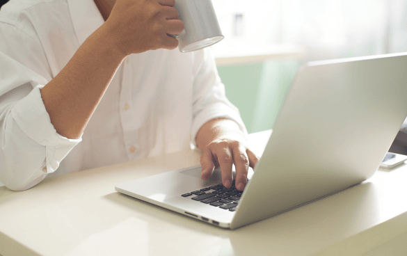 Person in a white shirt typing on a laptop while holding a white mug.