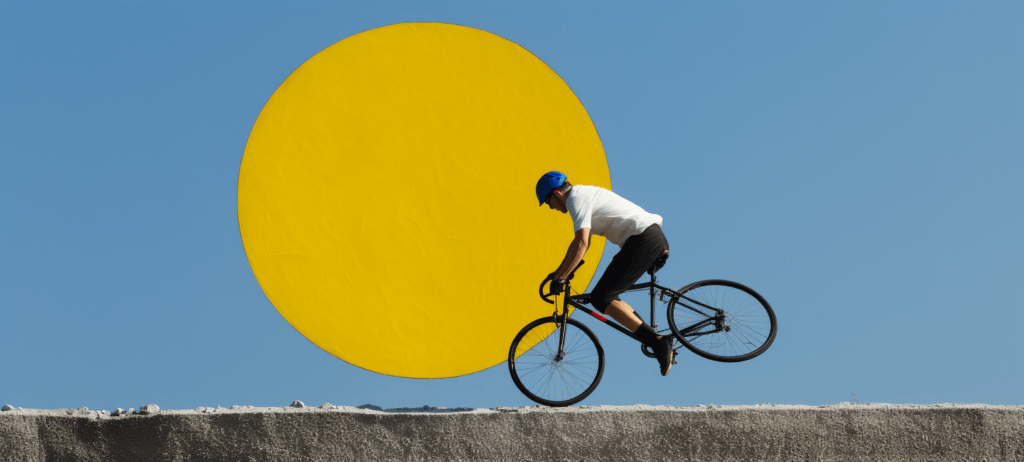 A person in a white shirt and blue helmet riding a bicycle on the edge of a concrete wall with a large yellow circle in the background, resembling a stylized sun against a blue sky.