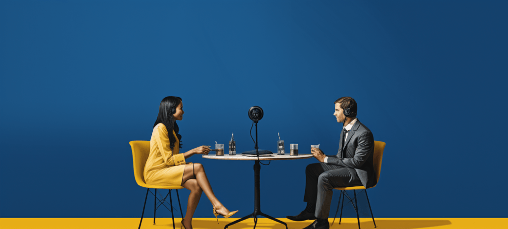 A woman in a yellow dress and a man in a suit sitting at a table with microphones and headphones, engaged in a podcast or interview against a blue background with a yellow floor strip.