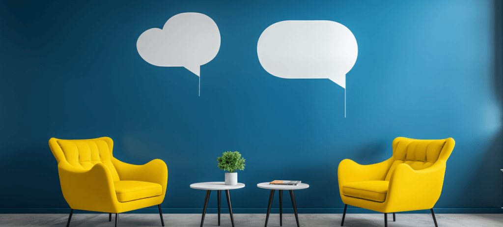 Two yellow armchairs facing each other with a white round table in between, on which there is a potted plant and reading materials, against a deep blue wall with white speech bubble decals, in a room with light wood flooring.