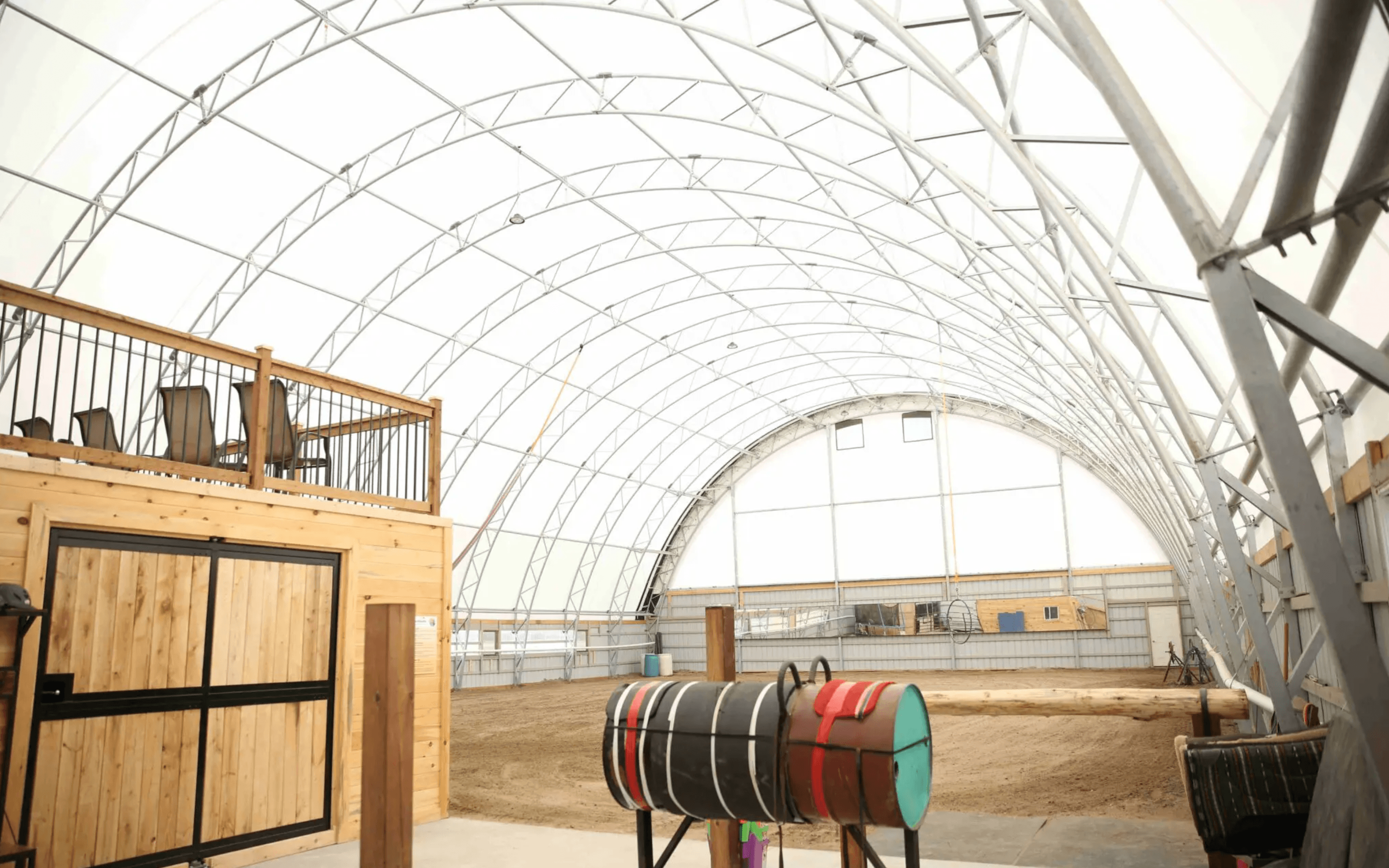 Indoor equestrian arena with a clear-span roof, sand-covered ground, and a wooden balcony with chairs. Colored barrels for horse exercises are visible in the foreground.