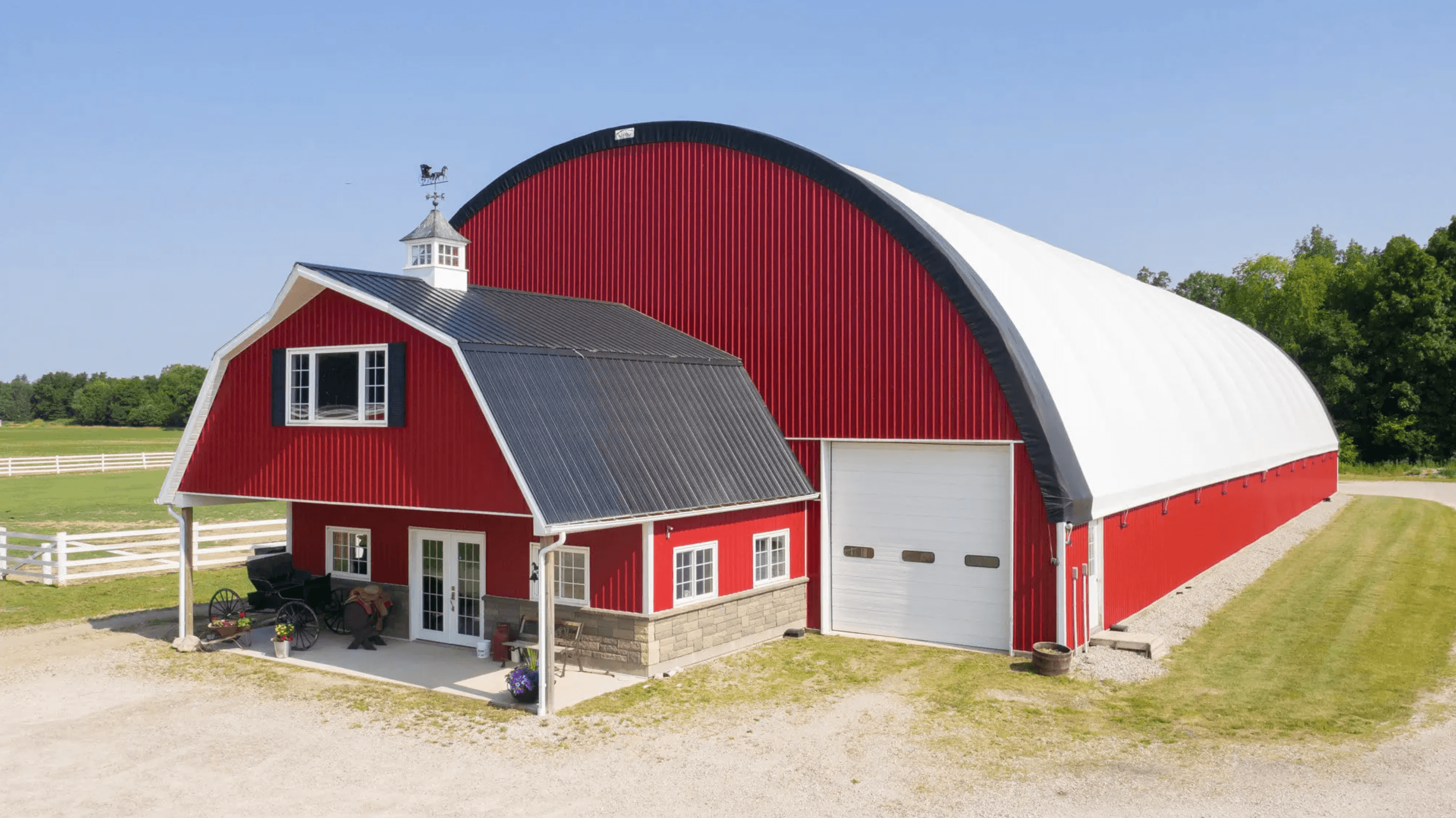 A large red barn with a white arched roof and attached smaller structure with a pitched roof, surrounded by a white fence, with a vintage carriage in front, set against a backdrop of greenery and a clear blue sky.